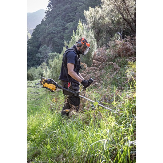 A person wearing protective gear and gloves uses a DEWALT DCMBC823N battery-powered brush cutter to trim dense grass and plants outdoors in a natural, hilly landscape.