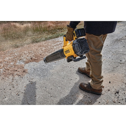 Person uses a DEWALT cordless blower to clear debris from a paved outdoor surface.