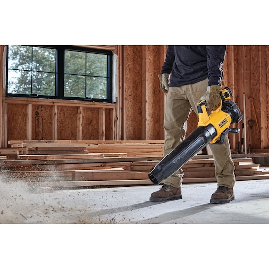 Person using a DEWALT cordless blower to clear dust in a wood-framed room under construction.