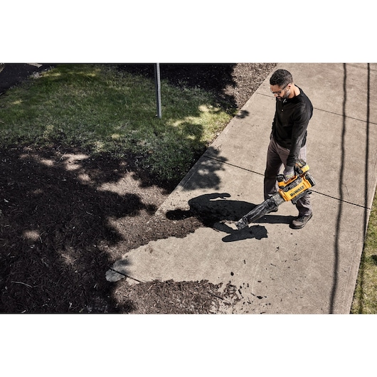 Person uses a DEWALT blower DCMBL777N to clear debris from a sidewalk outdoors.