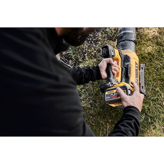 Person using a DEWALT cordless blower outdoors with a 54V FLEXVOLT battery, close-up on tool and hands.