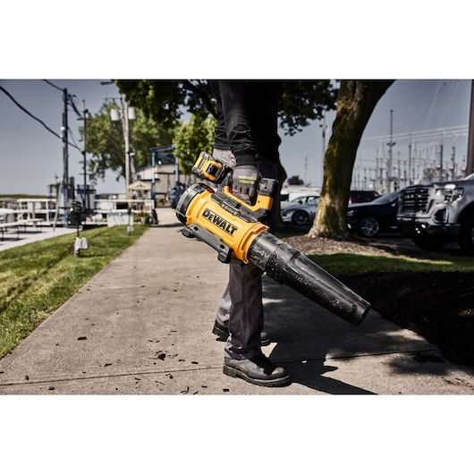 Person holding DEWALT cordless leaf blower while cleaning walkway outdoors near grass and parked cars.