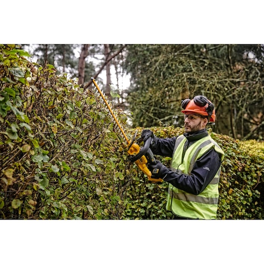 Person trimming hedge outdoors with a yellow DEWALT hedge trimmer, wearing safety gear and a reflective vest.