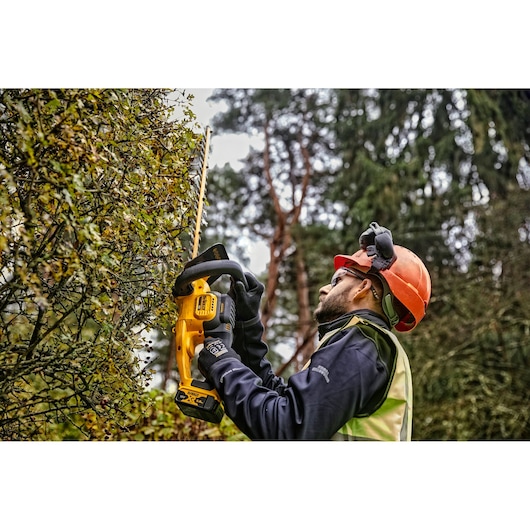 Person uses a DEWALT cordless hedge trimmer outdoors, wearing safety gear and gloves.