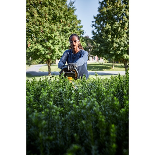 A person wearing gloves is using a DEWALT hedge trimmer to trim tall green bushes in a garden with trees and a clear sky in the background.