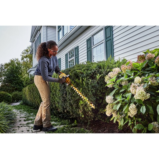 Person uses DEWALT cordless hedge trimmer to trim bushes in a garden outside a house.