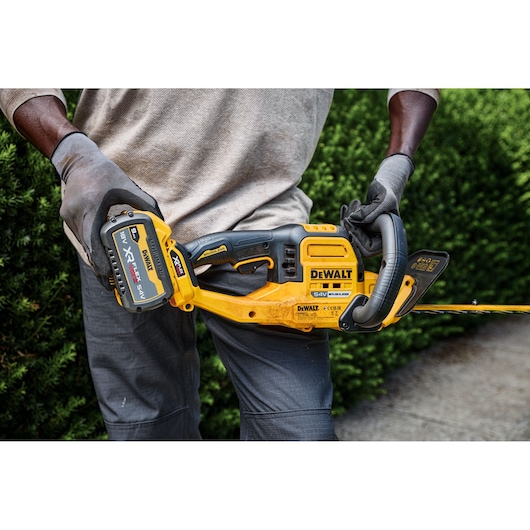 Person wearing gloves uses a DEWALT DCMHT573X1 cordless hedge trimmer outdoors near bushes.
