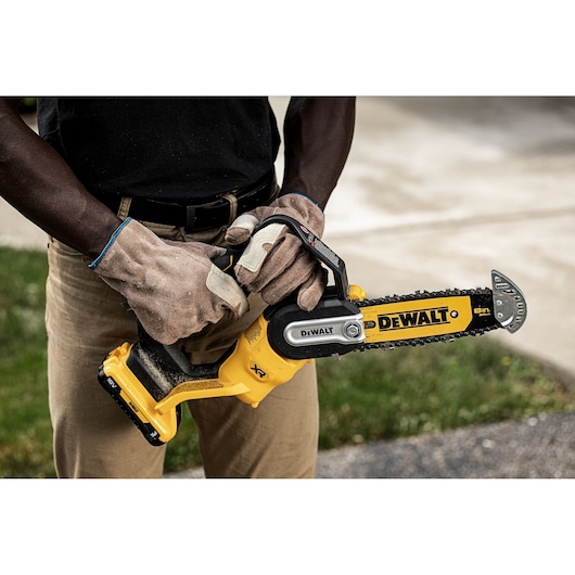 Person wearing gloves holds a DEWALT DCMPS520P1 cordless chainsaw outdoors.