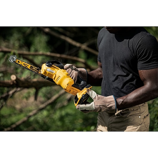 Person wearing gloves holds a DEWALT cordless chainsaw outdoors, preparing to cut tree branches.