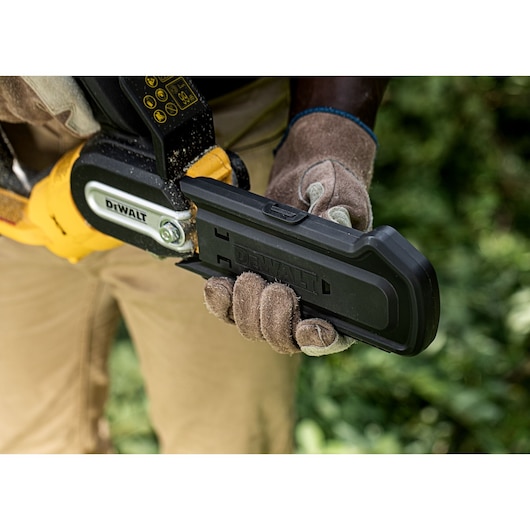 Gloved hand holds a DEWALT chainsaw with a protective blade cover outdoors.