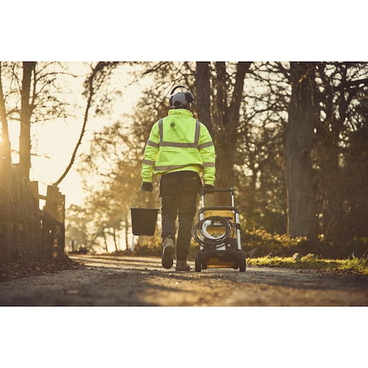 Person in safety gear pulls a DEWALT power washer and carries a bucket on a rural path at sunset.