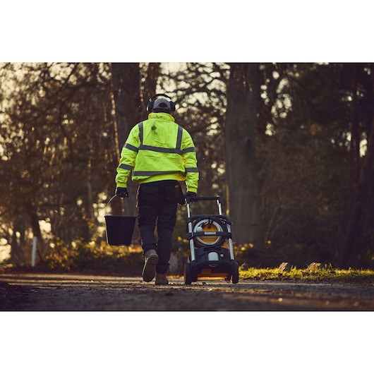 Worker in high-vis jacket pulls DEWALT pressure washer and carries bucket outdoors on path.