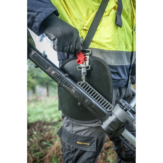 Close-up view of a person wearing protective clothing and gloves, holding a DEWALT brushless outdoor power tool with a harness, suitable for cutting or trimming vegetation.