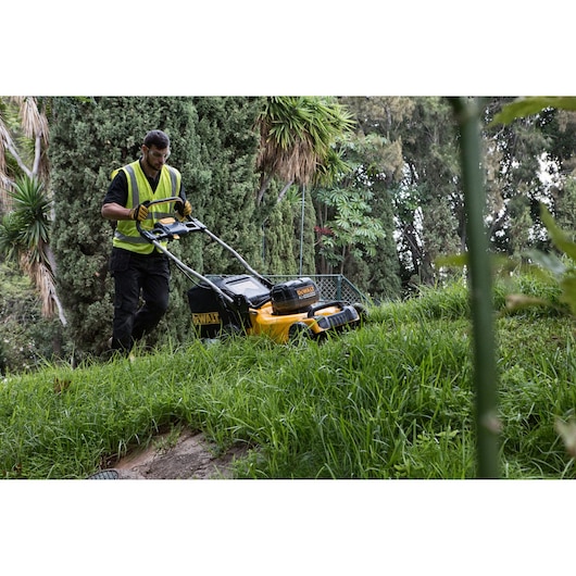 Person mowing grass with a DEWALT lawn mower on a slope, surrounded by trees and greenery.