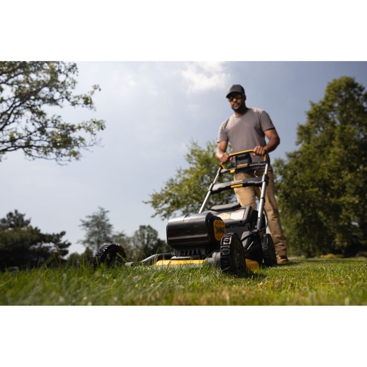 A person operates a DEWALT cordless lawn mower, model DCMWP134W2, outdoors on a grassy lawn with trees in the background. The person's face is blurred.