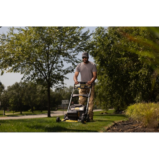 A person using a DEWALT cordless lawn mower (model DCMWP134W2) to cut grass in a garden or outdoor area, with trees and plants in the background.