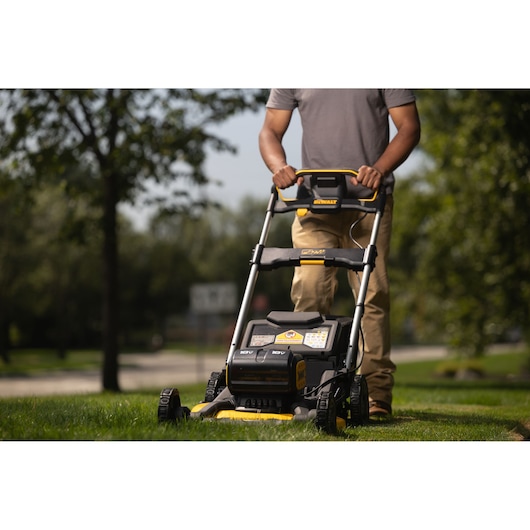 A person is pushing a DEWALT cordless lawn mower outdoors on a grassy lawn. The focus is on the lawn mower, showing its robust design and battery compartment.
