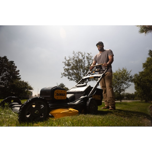 A person is pushing a DEWALT lawn mower (model DCMWP134W2) on a grassy area outdoors. The mower is black and yellow, and the person's face is blurred for privacy. There are trees and a cloudy sky in the background.