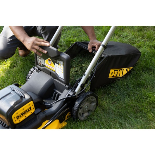 A close-up of a DEWALT cordless lawn mower on grass. A person is emptying the grass collection bag, which is attached to the mower.