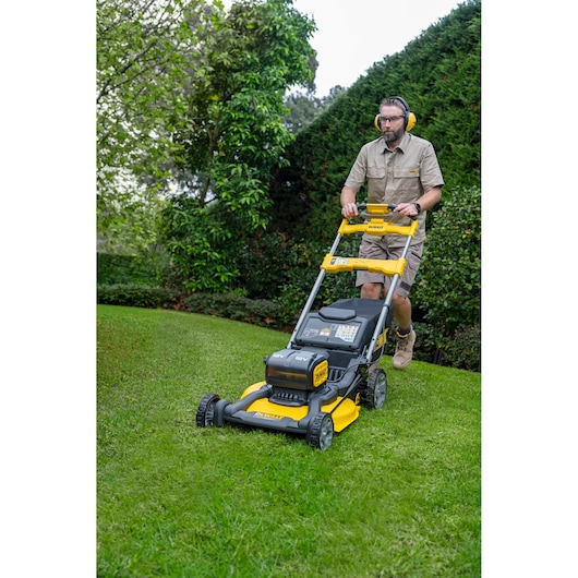 Person mowing lawn with DEWALT cordless lawn mower on green grass, wearing safety gear outdoors.