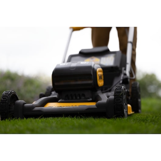 Close-up view of a DEWALT cordless lawn mower in use outdoors, showing the front wheels, mower deck, and battery