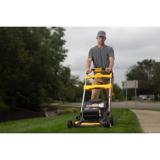 A person is pushing a yellow and black DEWALT lawn mower on green grass in a residential outdoor setting. The person's face