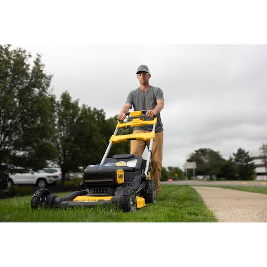 A person is using a DEWALT cordless lawn mower to cut grass along a sidewalk. The mower is black and yellow, and the