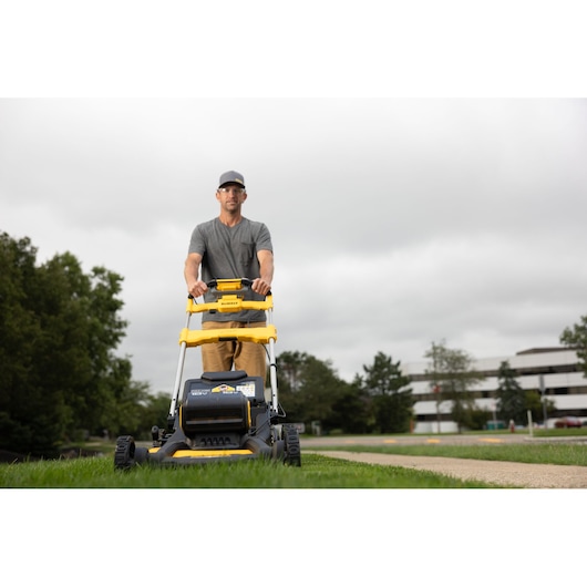 A person is pushing a DEWALT lawn mower on a grassy area outdoors. The image shows the mower from the front, and the