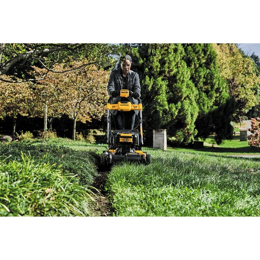 Person using a DEWALT lawn mower outdoors, cutting grass in a green garden with trees in the background.