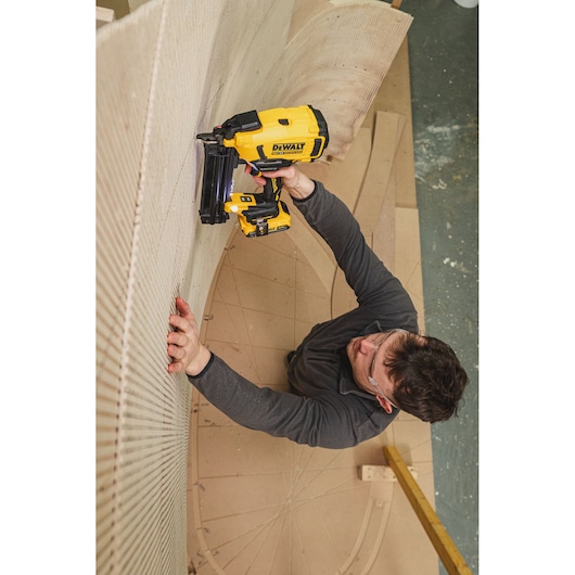 Person using DEWALT nailer DCN210SN-XJ on curved wood in workshop, wearing safety glasses and dark clothing.