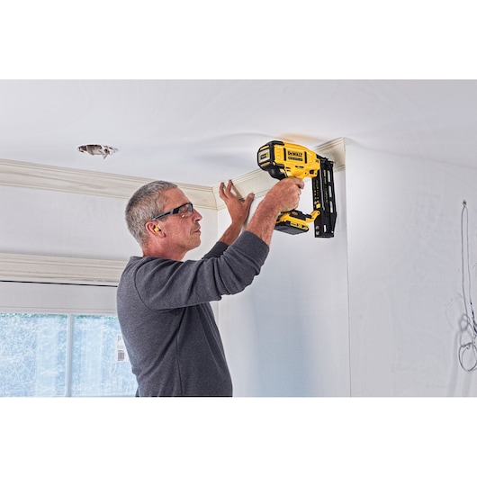 Person uses DEWALT DCN662N nailer to secure crown molding to a ceiling in a bright room.