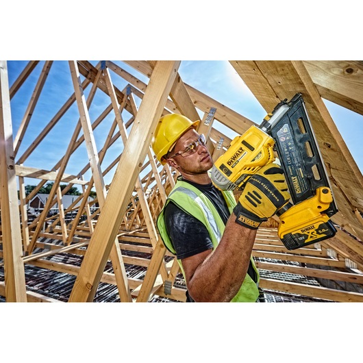Worker uses DEWALT DCN692N nailer on wooden beams at a construction site, wearing safety gear and gloves.