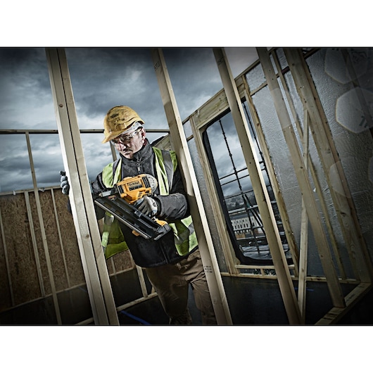Worker uses DEWALT nail gun DCN692P2 for wood framing at construction site, wearing safety gear.