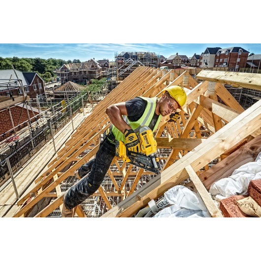 A construction worker wearing a yellow hard hat and high-visibility vest uses a DEWALT DCN692P2 nail gun to secure wooden beams on a house roof frame at an active building site. The surrounding area shows several houses under construction.