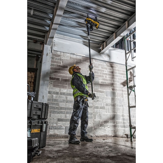 A construction worker wearing safety gear uses a DEWALT tool on an extension pole to work on a metal ceiling structure inside a building. DEWALT toolboxes are visible in the foreground.