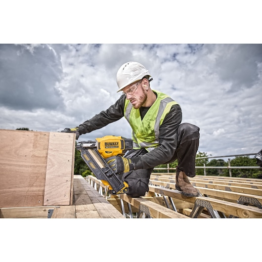 Person uses a DEWALT nail gun DCN930N on wooden boards at an outdoor construction site, wearing safety gear.