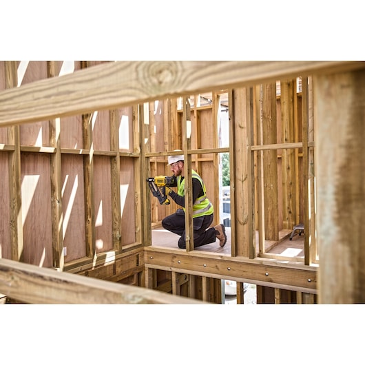 Worker uses a DEWALT tool to fasten wood frames at a construction site, wearing safety gear and kneeling.