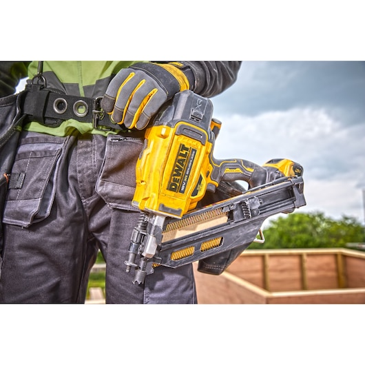 Person holding a DEWALT DCN930N nail gun, wearing work gloves and gear at a construction site outdoors.