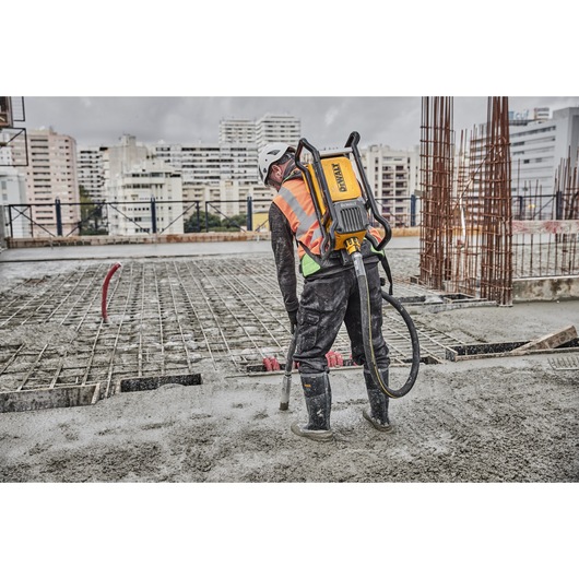 A construction worker wearing a safety helmet, high-visibility vest, and boots is using a DEWALT concrete vibrator on a large outdoor building site. The worker has the DEWALT device strapped to their back, and is standing on wet concrete near metal rebar structures, with city buildings visible in the background.