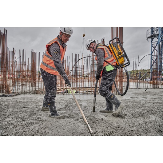 Two construction workers wearing safety helmets and orange reflective vests are working on a concrete slab at an outdoor construction site. One worker is using a DEWALT concrete vibrator backpack, while the other is spreading fresh concrete with a rake. Exposed rebar and industrial equipment are visible in the background.