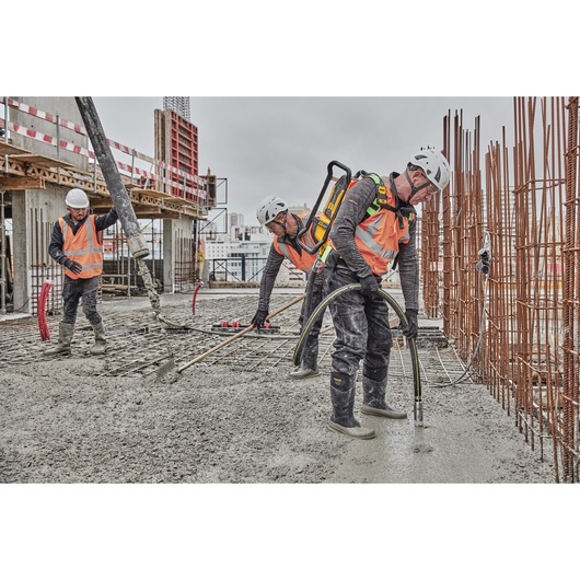 Construction workers wearing safety helmets and orange vests are pouring and leveling concrete on a building site. One worker uses a DEWALT cordless concrete vibrator, model DCPS310N, to help settle the concrete, while another handles a hose, and a third oversees the process. The site features steel reinforcement bars and partially built structures in the background.