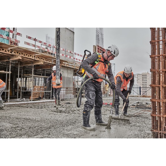 Construction workers wearing safety gear, including orange vests and helmets, use tools to spread and smooth concrete on a building site.
