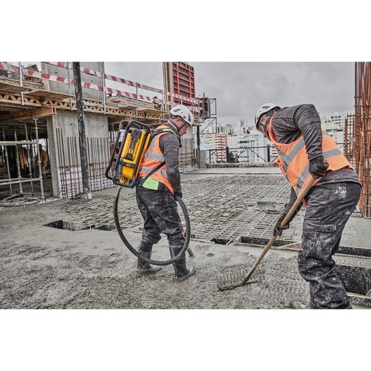 Two construction workers wearing orange safety vests and helmets are working on a concrete floor at a building site. One worker uses a DEWALT backpack-powered tool with a hose, while the other smooths the concrete with a rake.