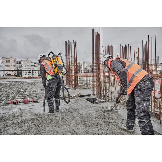 Two construction workers wearing safety helmets and orange reflective vests are working on a concrete surface at a building site. One worker uses a DEWALT backpack tool, while the other uses a rake to spread the concrete. Steel reinforcement bars are visible in the background.