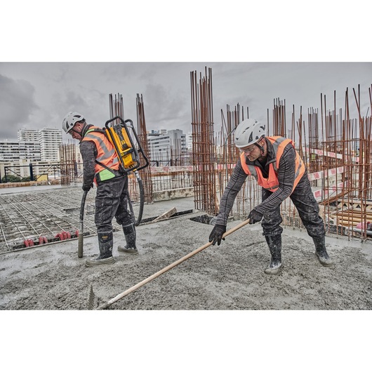 Two construction workers wearing safety helmets and orange vests are working on a concrete slab. One worker uses a DEWALT backpack tool to handle concrete while the other levels the wet concrete with a rake. Reinforcement bars and buildings are visible in the background.
