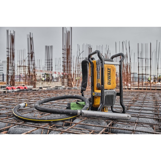 A DEWALT cordless concrete vibrator tool placed on a construction site with steel reinforcement bars and upright rebar columns. The tool is yellow with black accents and a flexible hose, suitable for professional construction work.
