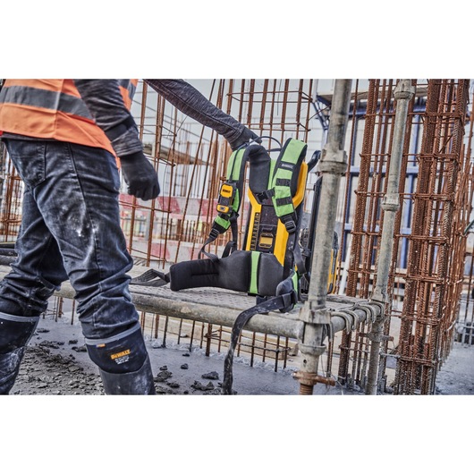 A DEWALT power tool harness with green and black straps is displayed on a metal construction site bench. A worker wearing a high-visibility orange vest, gloves, and protective gear stands nearby.