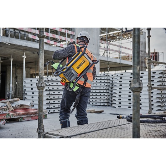 A construction worker wearing safety gear and a hard hat is carrying a DEWALT DCPS310N power tool on their back at a construction site.