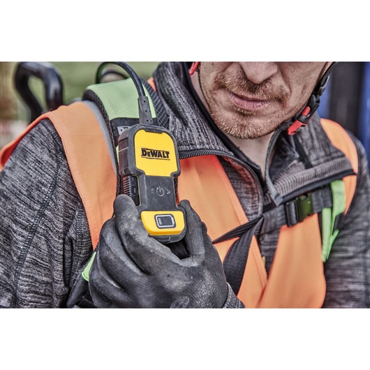 A person wearing an orange safety vest and black gloves holds a DEWALT DCPS310N device attached to their chest. The person's face is blurred for privacy.