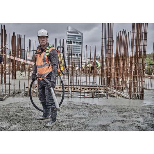 Person wearing a safety helmet and high-visibility vest, operating a DEWALT concrete vibrator tool (DCPS310N) at a construction site with rebar structures and wet concrete.